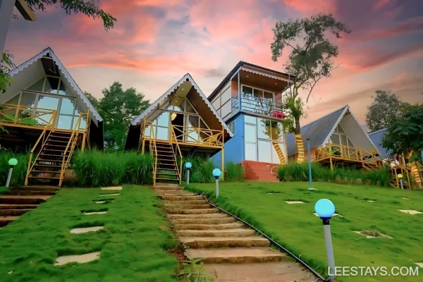 Scenic view of Diamond Resort by Pawna Lake, featuring unique cottages with wooden decks amidst lush greenery and a colorful sunset sky.