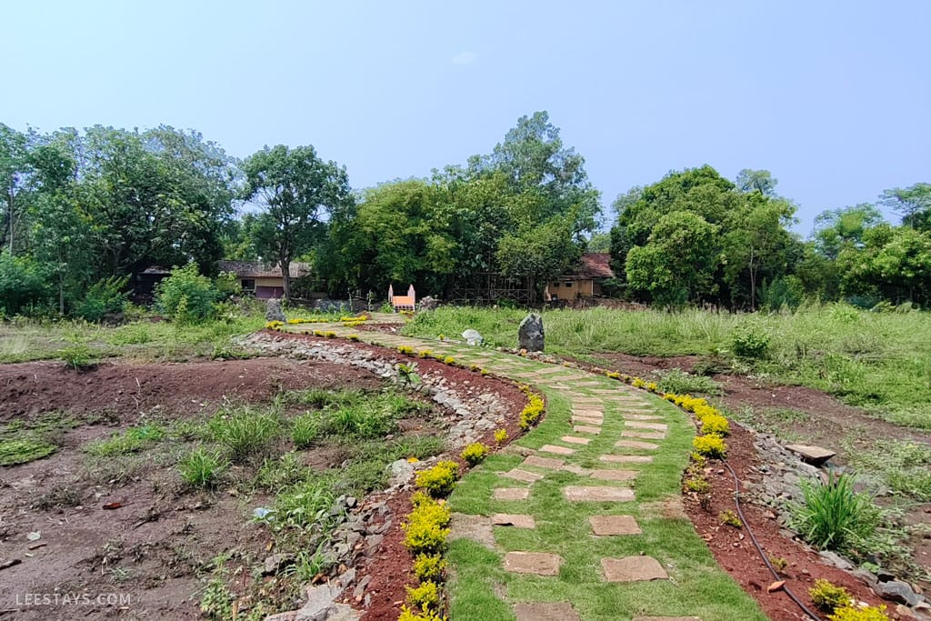 A scenic pathway lined with flowers and lush green grass, leading through a resort in Kamshet, surrounded by trees and open land.