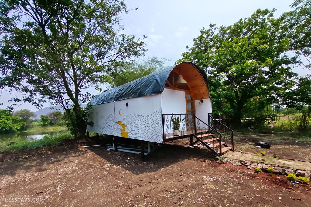 A unique accommodation structure at a resort in Kamshet, surrounded by trees and greenery, with a scenic view of a water body nearby.
