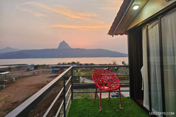 A scenic view of Pawna Lake from a balcony with a red chair, mountains in the background during sunset.