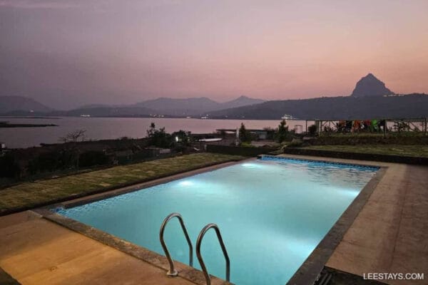 A serene view of Pawna Lake at dusk, featuring a pool and lush greenery under a colorful sky with mountains in the background.