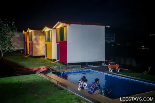 A couple sits by a pool outside colorful cabins at night, enjoying a relaxing stay by Pawna Lake.