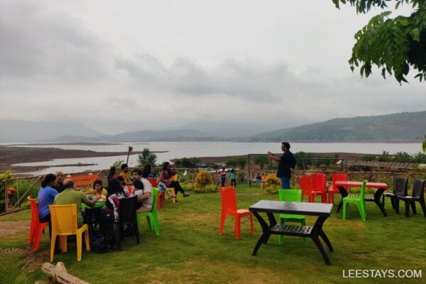 A vibrant outdoor seating area overlooking Pawna Lake with guests enjoying a meal, surrounded by lush greenery and cloudy skies.