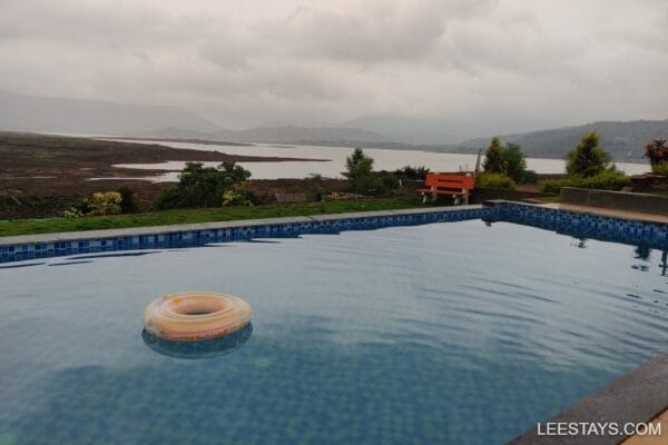 View of a serene pool at a stay near Pawna Lake, surrounded by lush greenery and distant hills under cloudy skies.