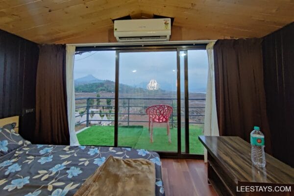 Cozy interior with a scenic view of Pawna Lake through large glass doors, featuring a bed, table, and a red chair on the balcony.