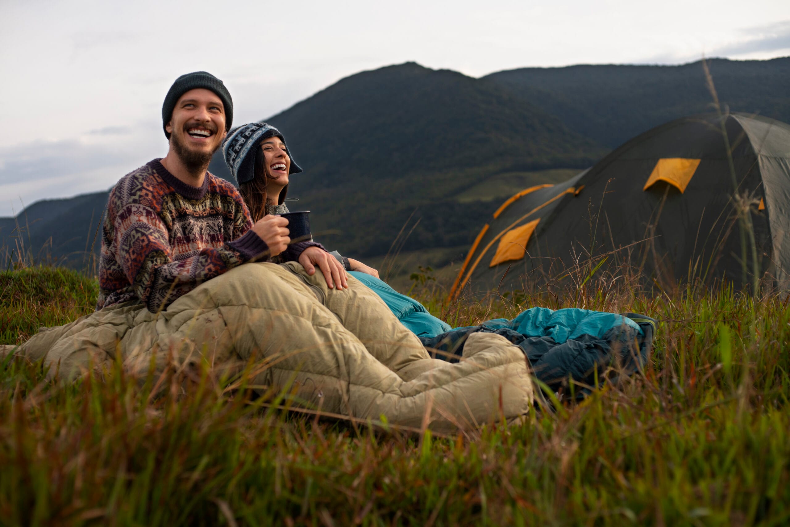 A smiling couple sitting on the grass wrapped in sleeping bags, enjoying a cup of coffee with mountains in the background and a tent nearby during a camping trip.