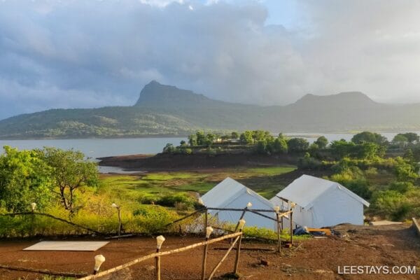Scenic view of glamping setups by Pawna Lake, surrounded by lush greenery and mountains under a cloudy sky.