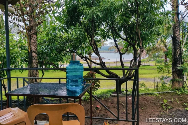 A serene view from a cottage balcony overlooking Pawna Lake, surrounded by lush greenery and a water container on the table.