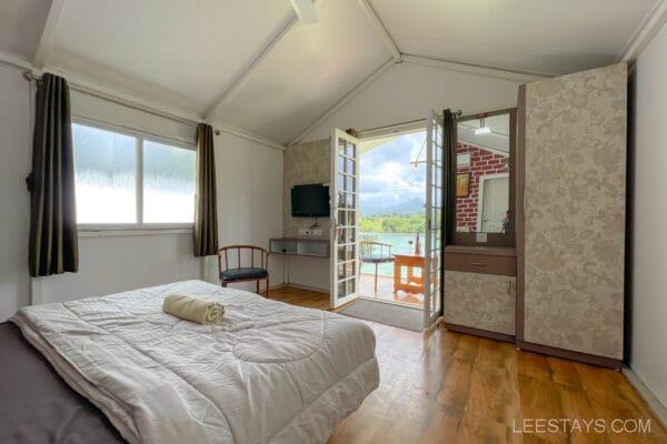 Cozy cottage interior with wooden flooring, a bed, and large windows overlooking the Pawna River, offering scenic views.