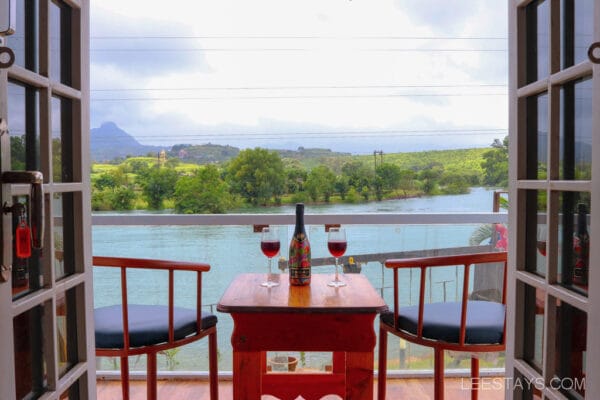 A cozy cottage overlooking the Pawna River, featuring a table with two glasses of wine and scenic greenery in the background.