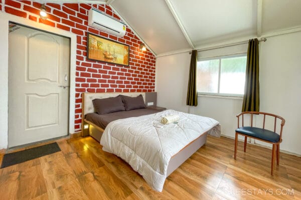 Cozy cottage interior overlooking the Pawna River, featuring a bed, decorative brick wall, and large windows with curtains.