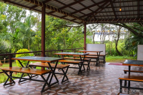 Outdoor dining area in a cottage overlooking the lush greenery by the Pawna River, featuring wooden tables and tiled flooring.