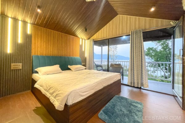 Modern cottage room with wooden interiors, a double bed, and large windows offering a view of Pawna Lake, ideal for a couple's getaway.