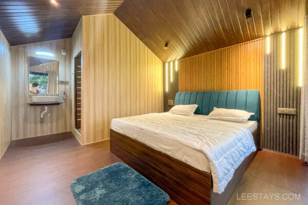 A cozy cottage interior by Pawna Lake, ideal for a couple, featuring a neatly made bed, wood-paneled walls, and a sink area with a mirror.