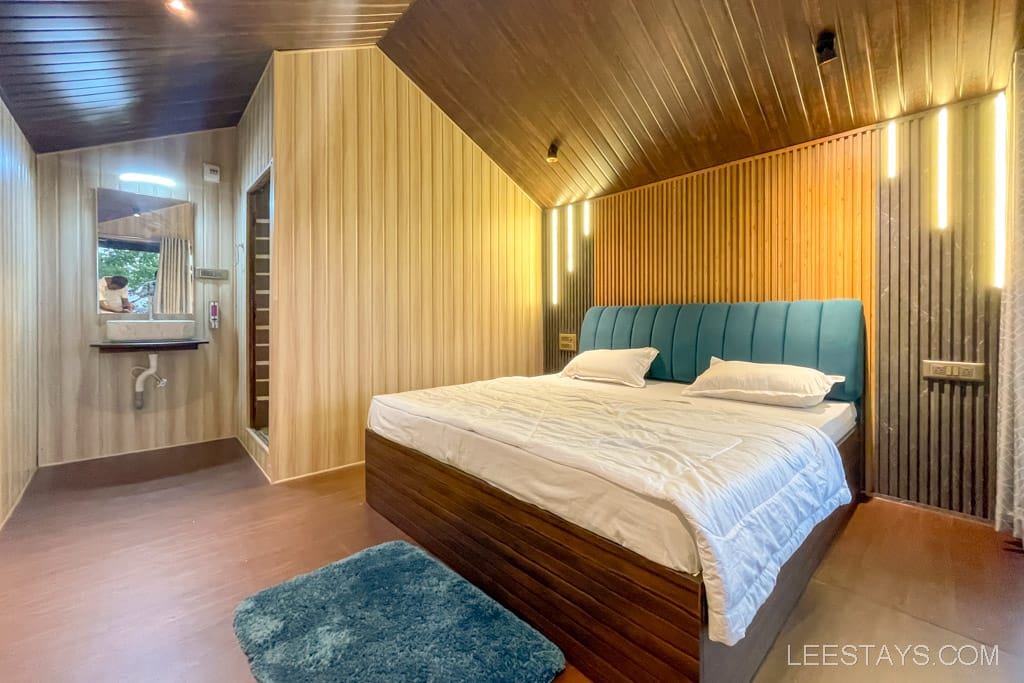 A cozy cottage interior by Pawna Lake, ideal for a couple, featuring a neatly made bed, wood-paneled walls, and a sink area with a mirror.