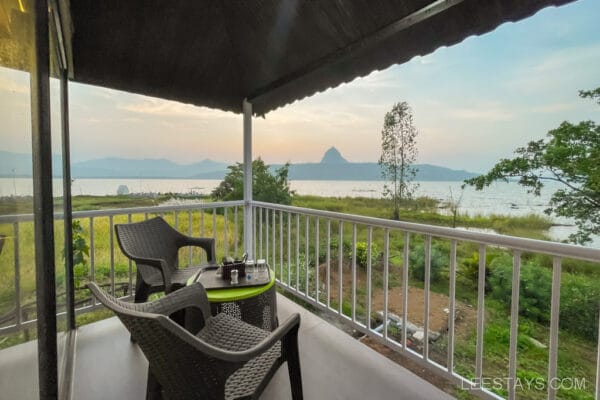 A scenic view of Pawna Lake from a cottage balcony with chairs and a table, perfect for a couple's getaway, surrounded by lush greenery and distant hills.