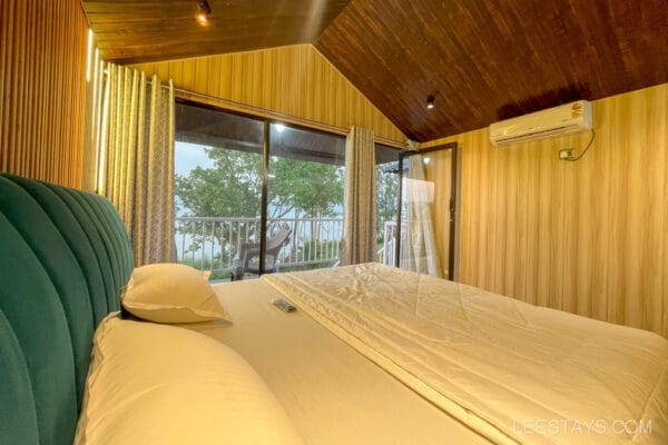 Cozy cottage interior for a couple, featuring a large bed with a view of trees by Pawna Lake visible through glass doors, offering a serene retreat.