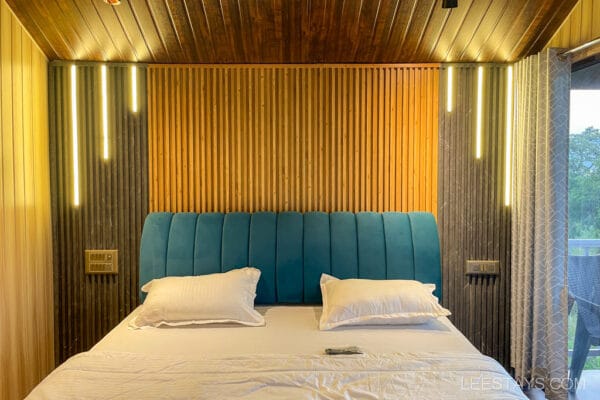 A cozy room ideal for a couple in a cottage by Pawna Lake, featuring a bed with a blue velvet headboard and warmly lit wood-paneled walls.