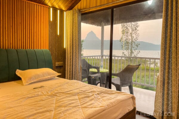 Cozy cottage bedroom with a view of Pawna Lake, ideal for a couple, featuring a neatly made bed and a small balcony with chairs overlooking the water.