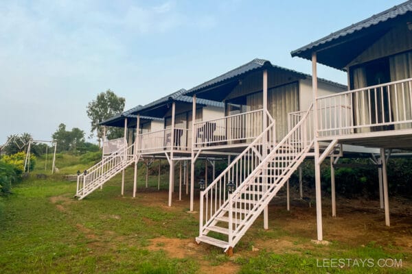A row of elevated cottages near Pawna Lake, suitable for couples, with wooden stairs and surrounded by greenery and trees under a clear sky.