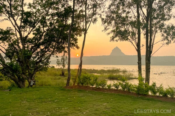 Scenic view of Pawna Lake at sunset with a grassy area, ideal for couples, surrounded by trees and distant cottages near the lakeshore.