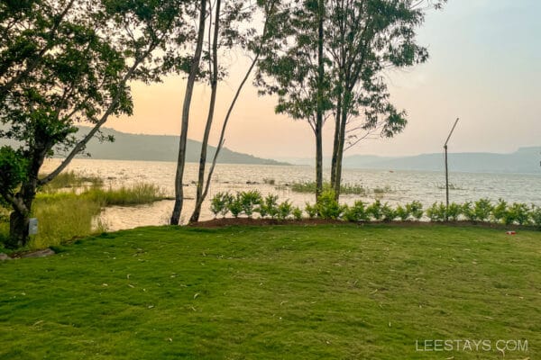 A serene view of Pawna Lake with lush green lawn and trees, ideal for couples visiting cottages nearby for a relaxing getaway.