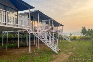 A series of elevated cottages near Pawna Lake, ideal for couples, with stairs leading up to each verandah, against a backdrop of tranquil water and sunset.