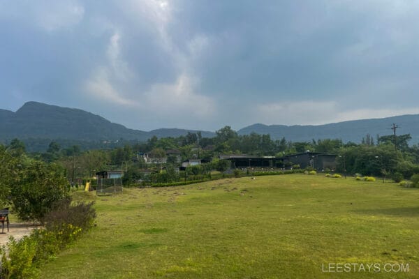 View of a resort near Pawna Lake with green lawns, surrounding trees, and distant hills under a cloudy sky.
