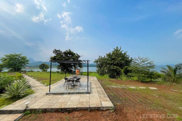 Outdoor dining area with a table and chairs overlooking scenic Pawna Lake at a resort, surrounded by greenery and a clear blue sky.