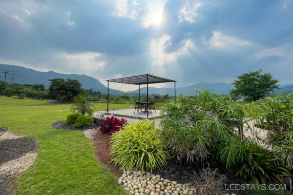 A serene view of a resort near Pawna Lake, featuring a pergola with outdoor seating amidst lush green grass and plants, under a dramatic cloudy sky.