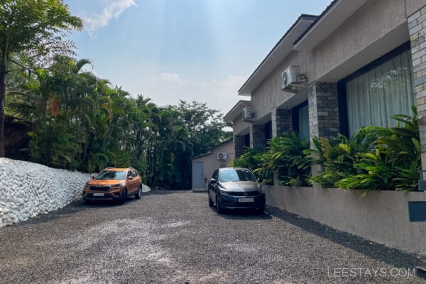 Driveway at a resort near Pawna Lake, featuring two parked cars, lush greenery, and a modern building exterior on a sunny day.