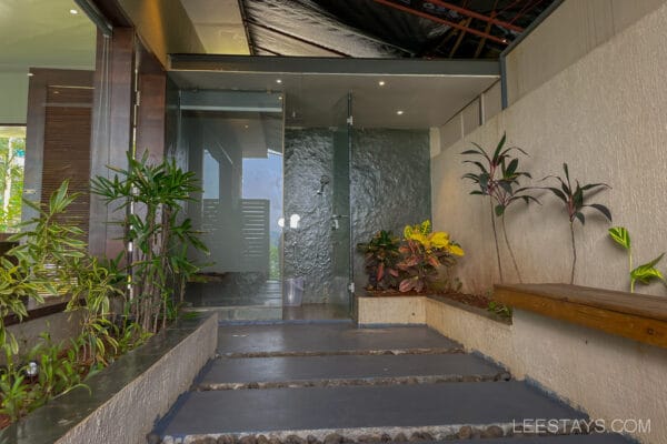 Outdoor shower area with glass enclosure and lush plants at a resort near Pawna Lake, featuring modern architectural design and natural elements.
