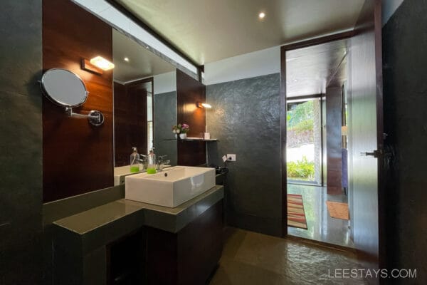 Modern bathroom with sleek design at a resort near Pawna Lake, featuring a large mirror, countertop sink, and view of greenery through the window.