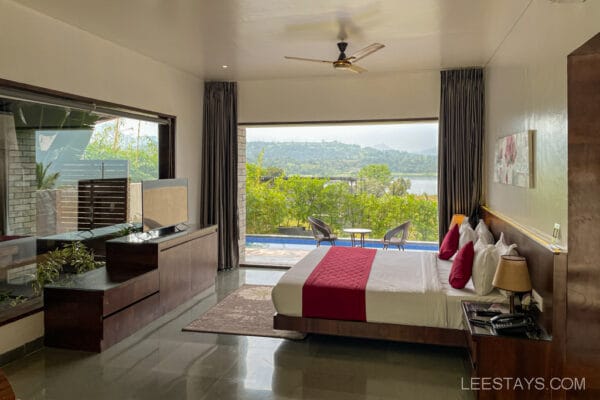 Luxurious resort room near Pawna Lake, featuring a bed with red accents, TV, and large window with stunning lake and landscape view.