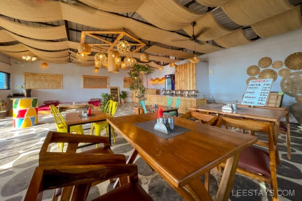 A cozy dining area with wooden tables and colorful chairs, part of glass rooms near Pawna Lake with decorative hanging lights and woven ceiling decor.