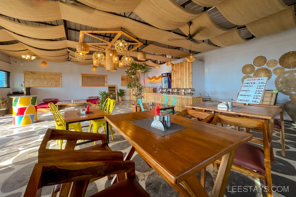 A cozy dining area with wooden tables and colorful chairs, part of glass rooms near Pawna Lake with decorative hanging lights and woven ceiling decor.