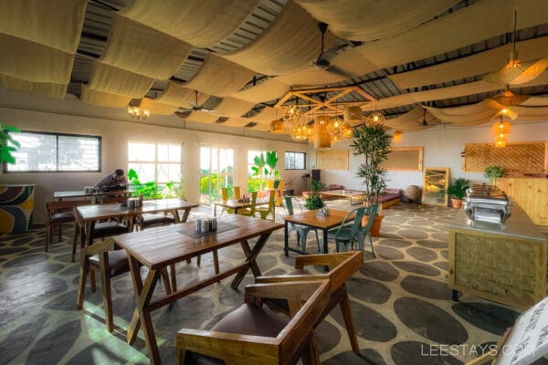 Spacious glass-walled room near Pawna Lake with wooden furniture, potted plants, and warm lighting under a decorative ceiling.