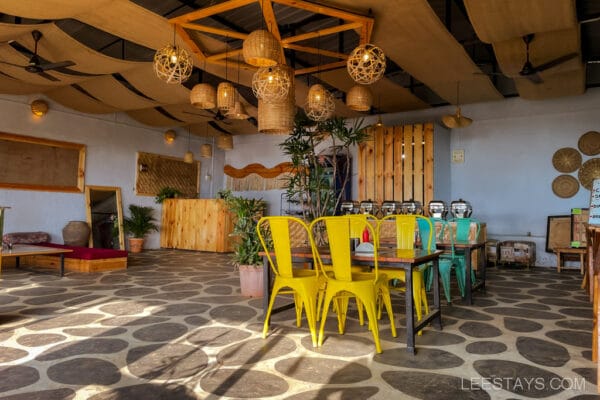 Interior of a glass room near Pawna Lake featuring colorful chairs, woven light fixtures, and decorative plants under a wooden ceiling.