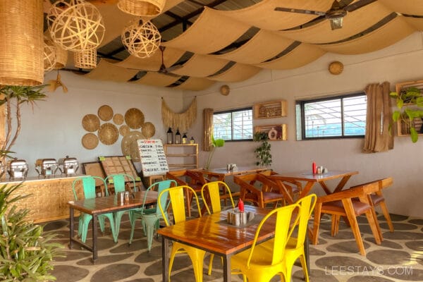 A cozy dining area in Pawna Lake features colorful chairs and tables, woven decor, and glass partitions; perfect for enjoying the glass rooms vibe.