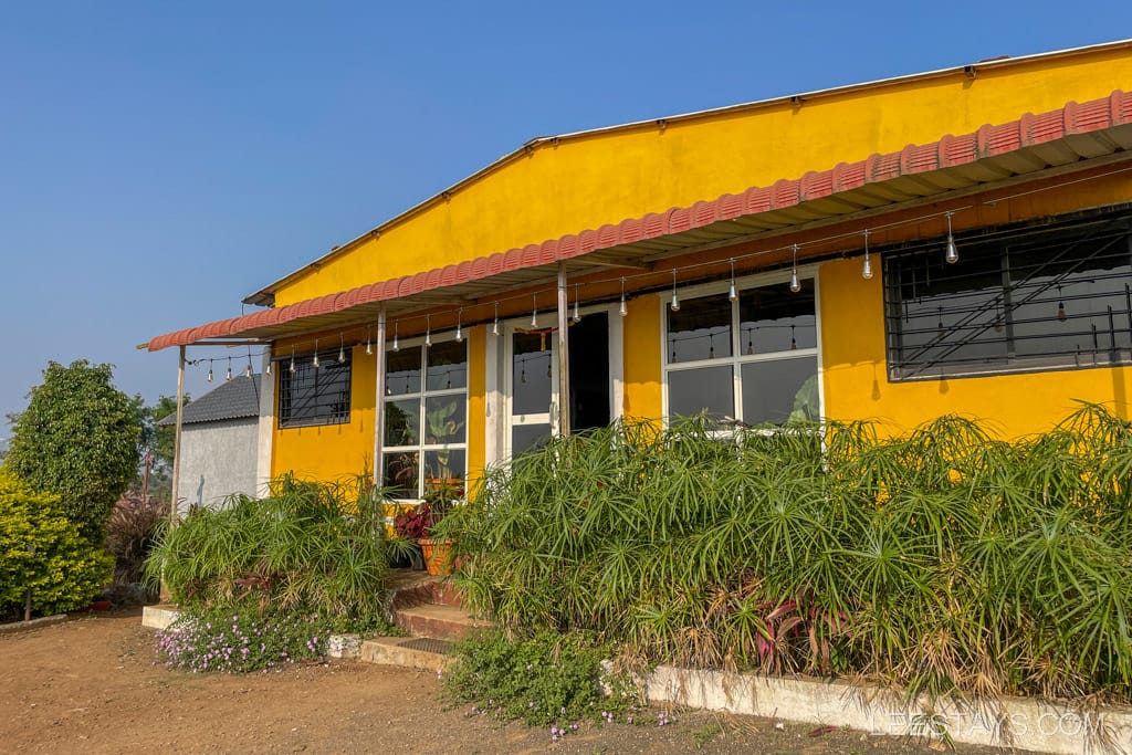 Yellow building with large glass windows known as "glass rooms" located near Pawna Lake, surrounded by greenery and clear blue skies.