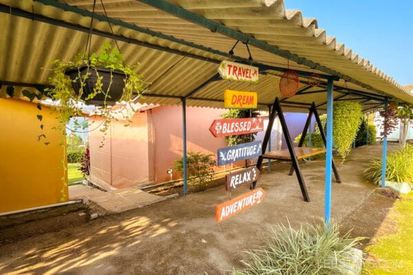 Outdoor area with vibrant signs for travel and relaxation, near glass rooms at Pawna Lake, under a corrugated roof with hanging plants.