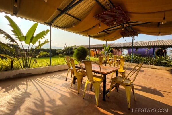 Outdoor dining area with yellow chairs and wooden table, shaded by a canopy, near glass rooms with views of Pawna Lake.