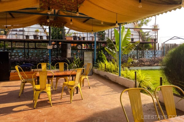 Outdoor seating area with yellow chairs under a canopy, in view of garden and glass rooms near Pawna Lake.