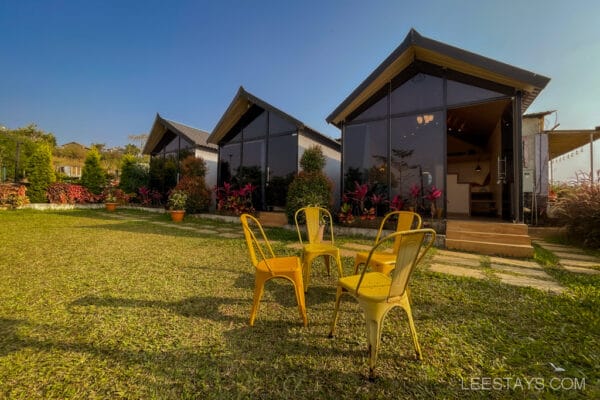 Glass rooms near Pawna Lake with yellow chairs on a grassy lawn, surrounded by greenery and clear blue sky overhead.
