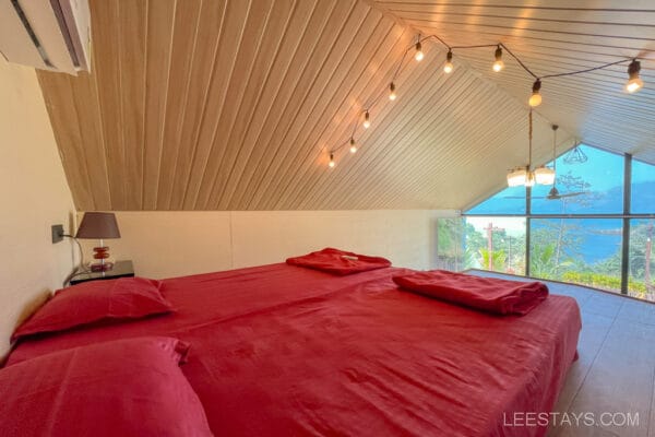 A cozy bedroom with red bedding, situated in a glass room overlooking Pawna Lake, featuring a sloped wooden ceiling and hanging string lights.