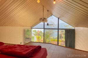 A cozy glass room overlooking Pawna Lake, featuring a wooden ceiling, bright natural light, and a bed with red bedding.