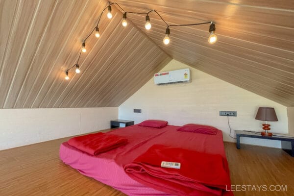 Cozy attic bedroom with a sloped wooden ceiling, string lights, and red bedding, resembling accommodations in glass rooms near Pawna Lake.
