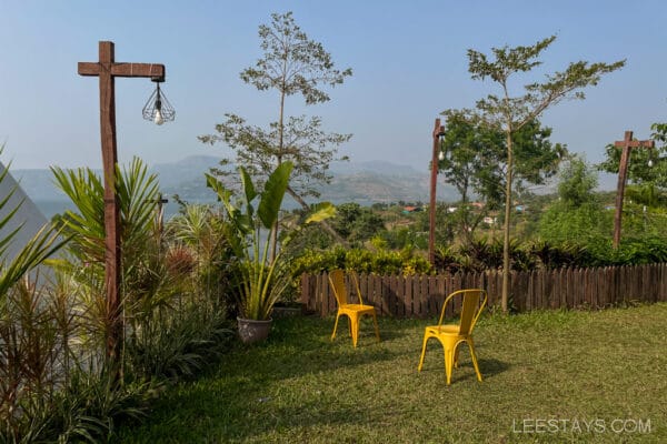 Two yellow chairs on a grassy area overlooking Pawna Lake, with a garden and glass rooms nearby, surrounded by lush greenery and distant hills.