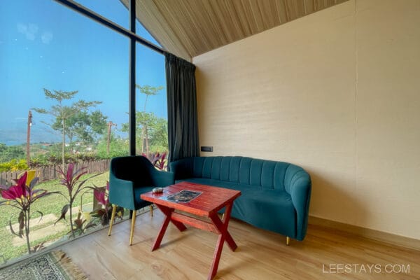 Interior of a glass room near Pawna Lake featuring a teal sofa, black chair, and wooden table with a large window overlooking green scenery.