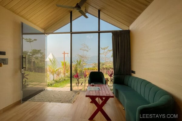 Interior of a cozy glass room with a view of Pawna Lake through large windows, green sofa, and wooden table, blending nature and comfort.